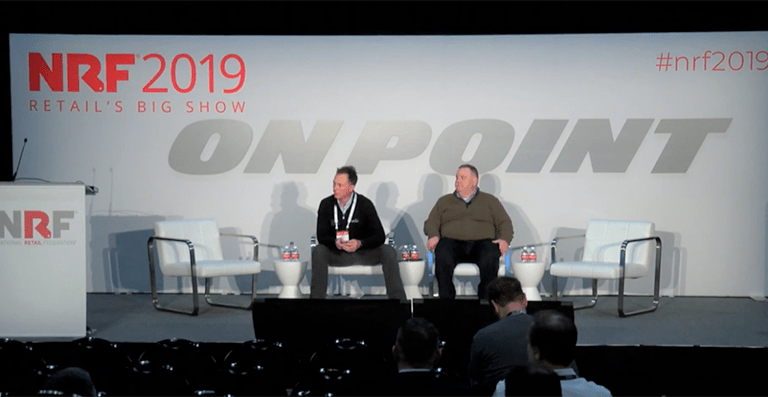 Two men sit on a stage with three empty chairs at NRF 2019 Retail’s Big Show. The backdrop has “ON POINT” and event branding. A podium is on the left, and a few people are seated in the audience.