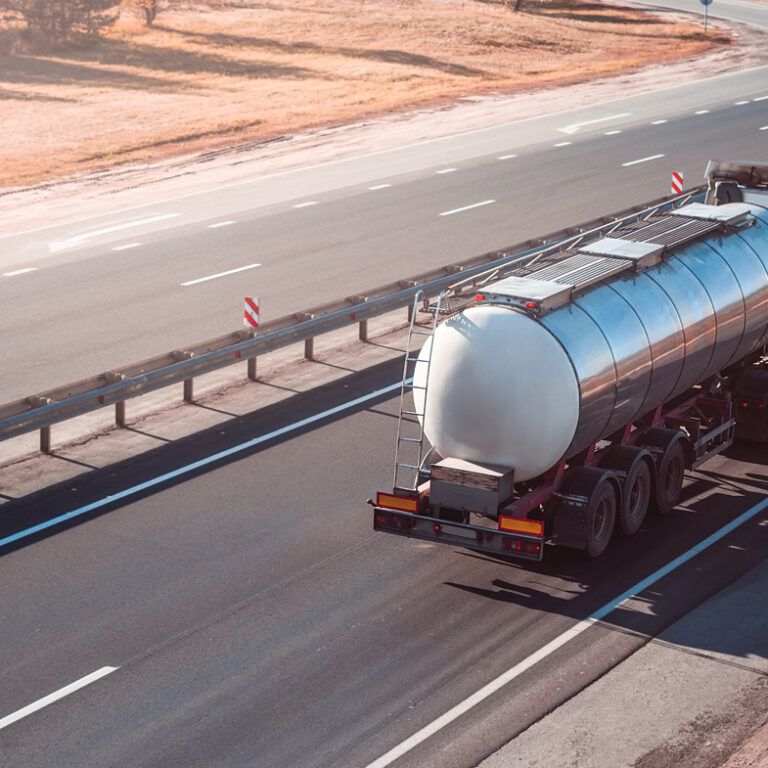 A silver tanker truck drives along an empty multi-lane highway on a clear day, with dry grass visible on the roadside.
