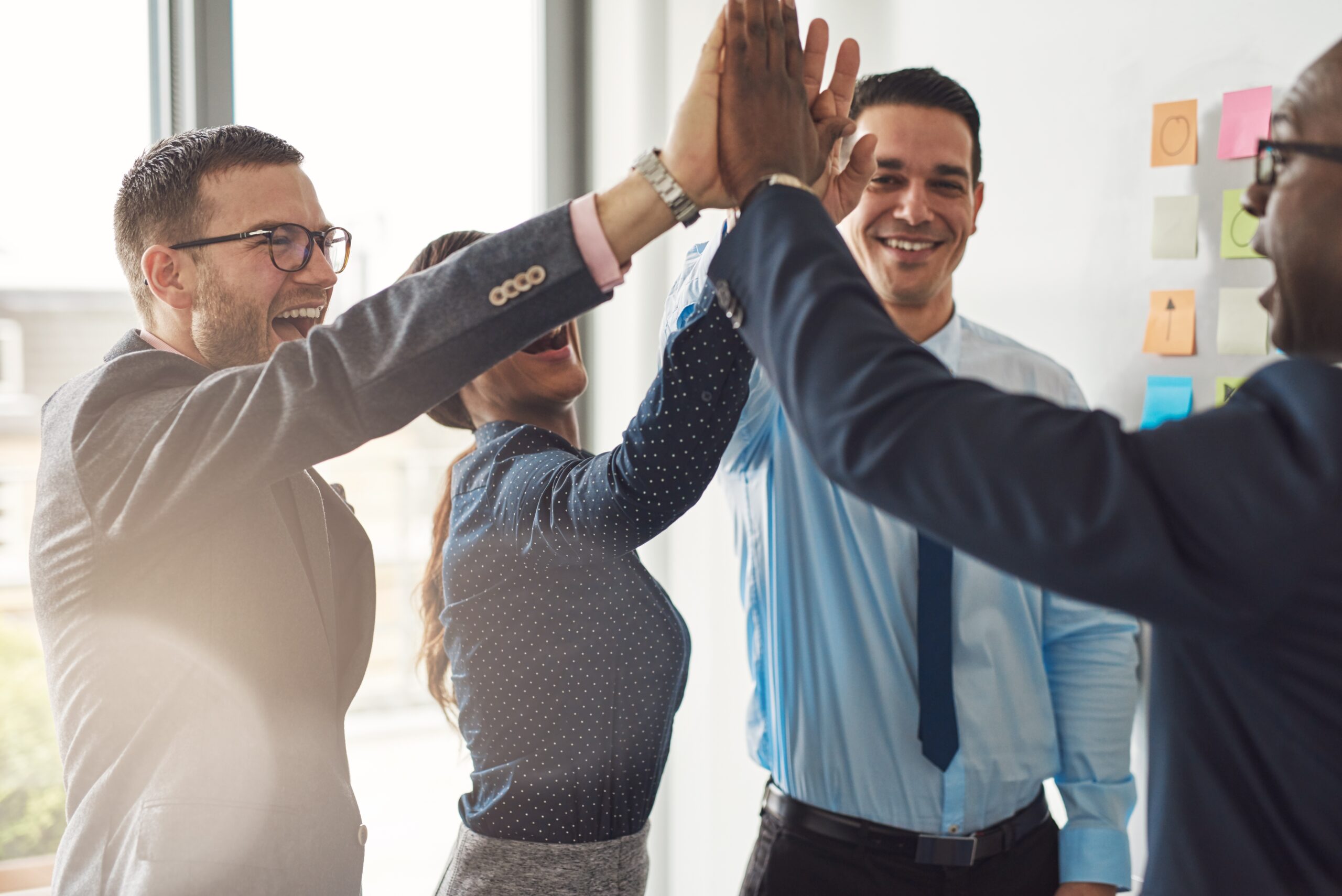 Four business professionals, three men and one woman, smiling and giving a group high-five in an office setting with sticky notes on the wall in the background.