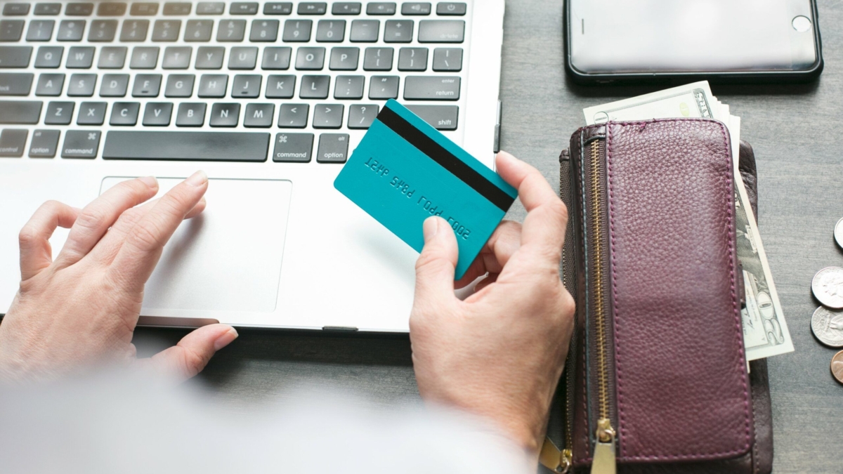 A person uses a laptop while holding a blue credit card; a purple wallet with cash, coins, and a smartphone are nearby on the table.