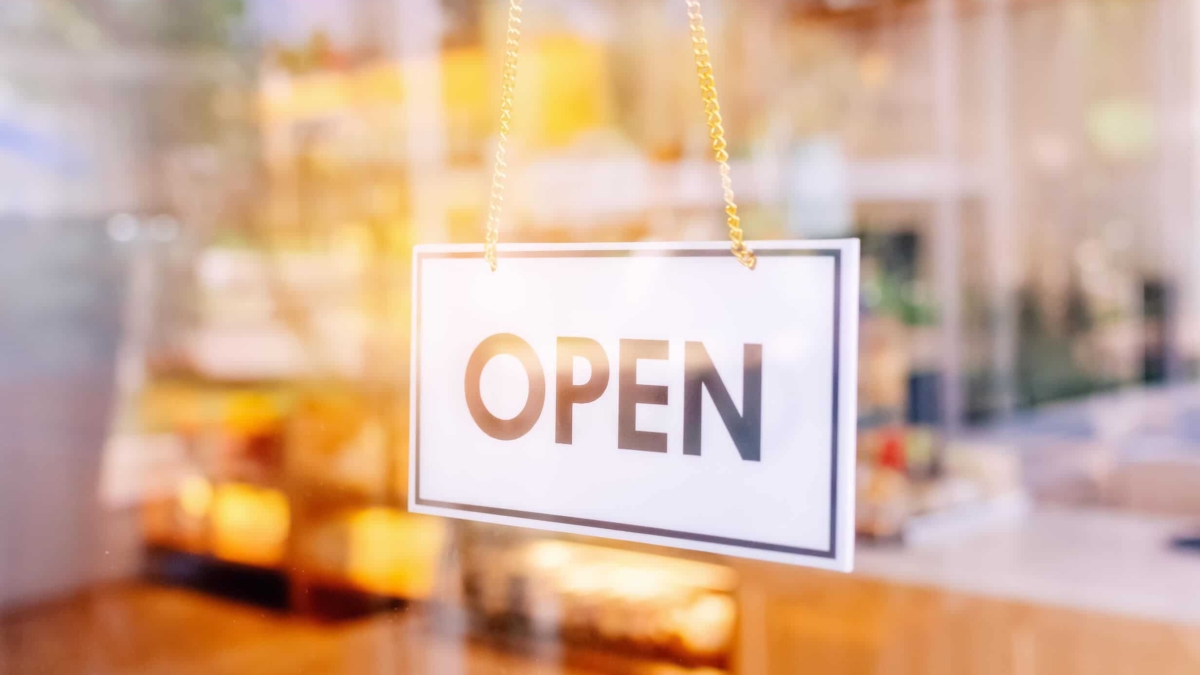 A white sign with the word OPEN in bold black letters hangs on a glass door, with a blurred view of a brightly lit interior in the background.