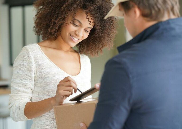 A woman smiles as she receives a package from a delivery person and signs for it on a digital device.