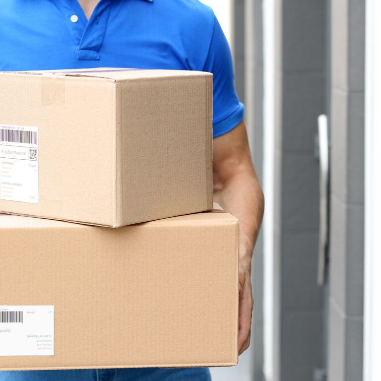 A person wearing a blue polo shirt is holding two cardboard packages with shipping labels, standing outside in front of a building with a gray wall and door.