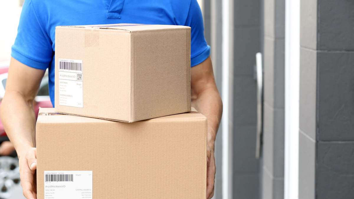 A person wearing a blue polo shirt is holding two cardboard packages with shipping labels, standing outside in front of a building with a gray wall and door.