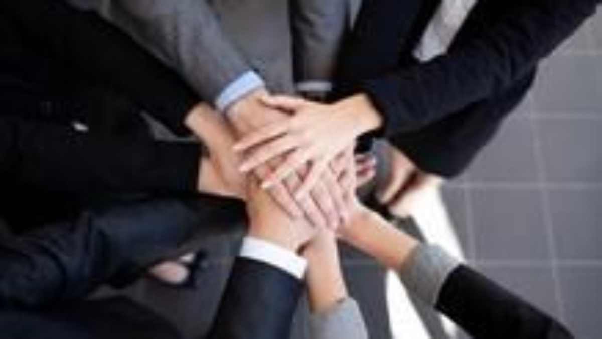 A group of people standing in a circle place their hands together in the center, symbolizing teamwork and unity. All are dressed in business attire.
