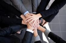 A group of people standing in a circle place their hands together in the center, symbolizing teamwork and unity. All are dressed in business attire.