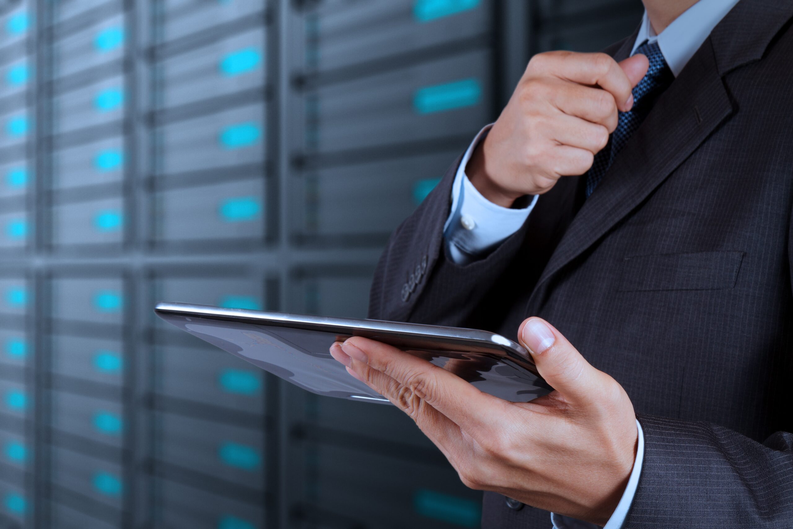 A person in a suit adjusts their tie while holding a tablet, standing in front of a blurred background of server racks in a data center.