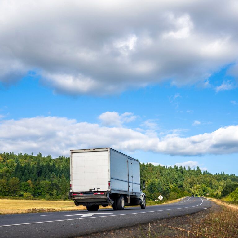 A white truck drives on a winding rural road bordered by green trees and fields, with a blue sky and scattered clouds overhead.