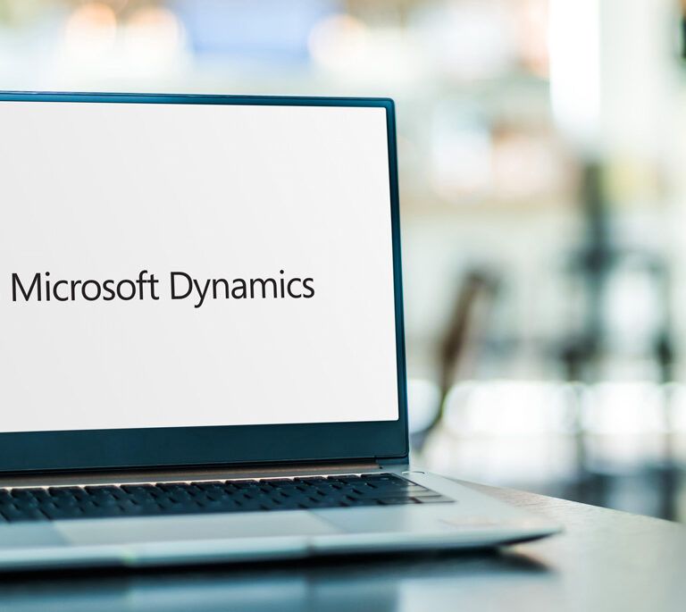 A laptop on a table displays the Microsoft Dynamics logo on its screen. The background is blurred, suggesting an office or workspace environment.