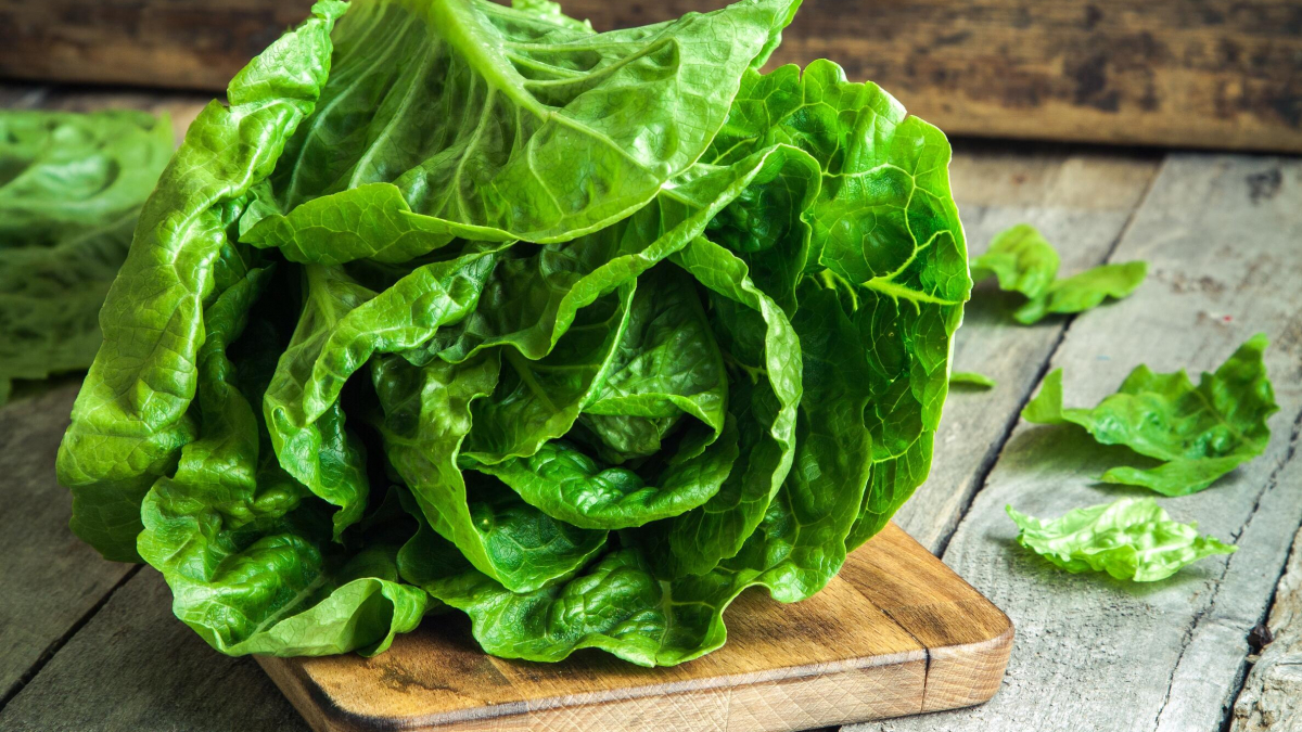 A fresh head of green lettuce sits on a small wooden cutting board, with a few loose lettuce leaves nearby, on a rustic wooden surface.