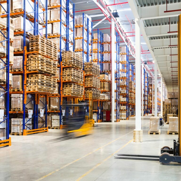 Warehouse with tall shelves stacked with boxes and pallets, a blurred forklift moving goods down the aisle, and another yellow forklift parked in the foreground; a bright and organized environment shows what slotting can achieve.