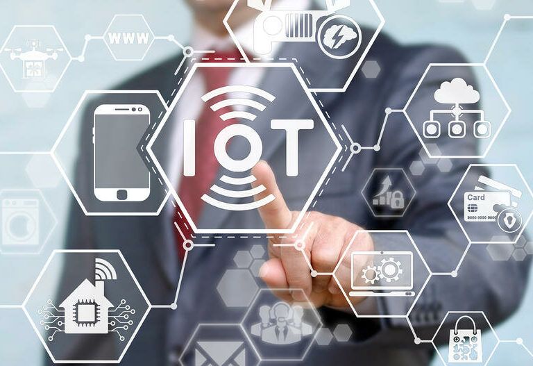 A person in a suit touches a virtual touchscreen with the word IOT at the center, surrounded by hexagonal icons representing technology and Internet of Things devices, such as a phone, house, and car.