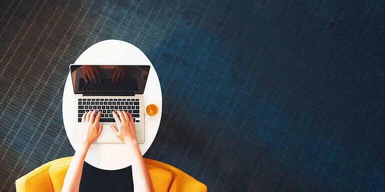 Top-down view of a person typing on a laptop at a small white table, with a cup of coffee nearby. The person is seated in a yellow chair on a dark blue textured floor.