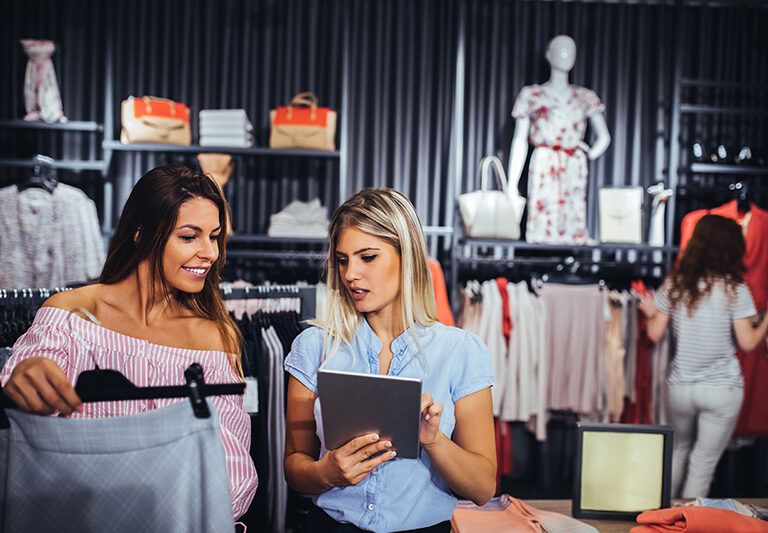 Two women stand in a clothing store, one holding a skirt and the other using a tablet. Clothing racks and accessories are visible, with another person browsing in the background. Mannequins display outfits.