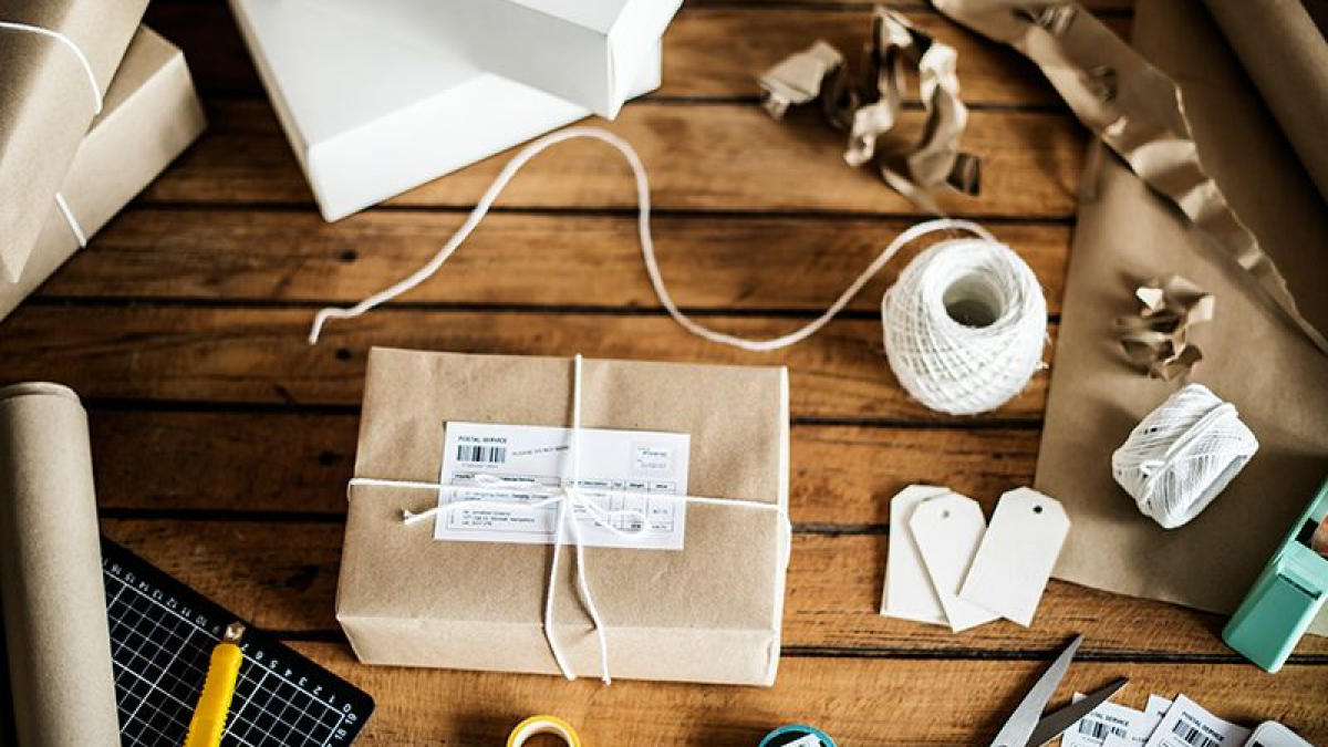 A wooden table with a parcel wrapped in brown paper and white string, surrounded by packing materials, scissors, shipping labels, blank tags, a utility knife, a ruler, tape, and white boxes.
