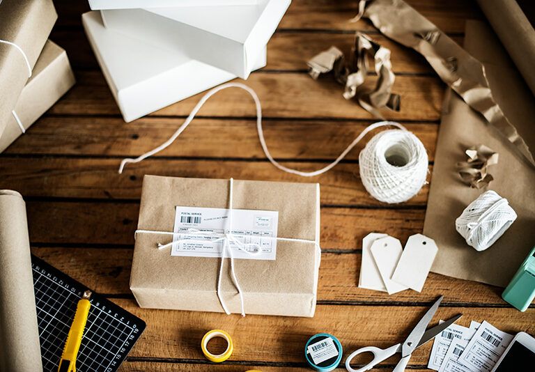 A wooden table with a parcel wrapped in brown paper and white string, surrounded by packing materials, scissors, shipping labels, blank tags, a utility knife, a ruler, tape, and white boxes.