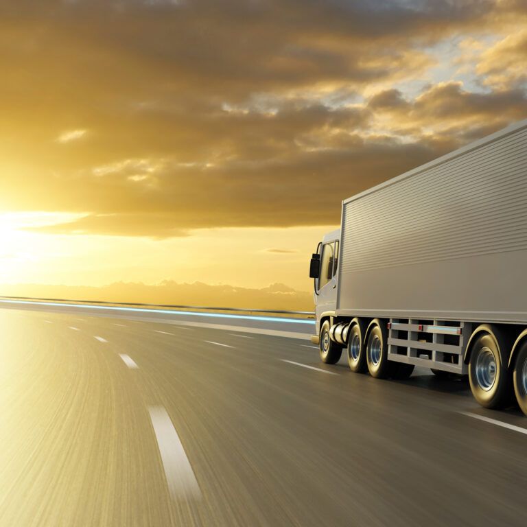 A large white semi-truck drives down an empty highway at sunset, with golden sunlight illuminating the road and dramatic clouds in the sky.