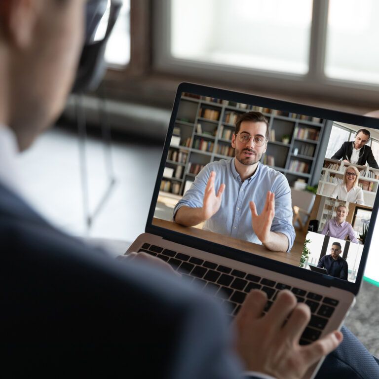 A person in a suit participates in a video conference on a laptop, showing five people on screen, each in separate windows, engaged in discussion. A potted plant and large windows are visible in the background.