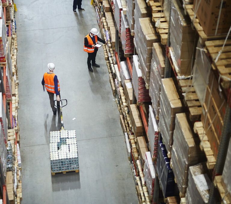 Workers in orange vests and helmets move among tall shelves stacked with boxes in a large warehouse. One worker uses a pallet jack to move goods, while another checks inventory with a clipboard.
