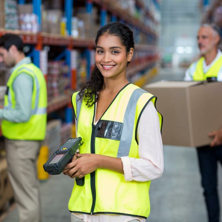 Smiling woman in a yellow safety vest holds a barcode scanner in a warehouse, while two coworkers organize and carry boxes in the background. Shelves with various items line both sides.