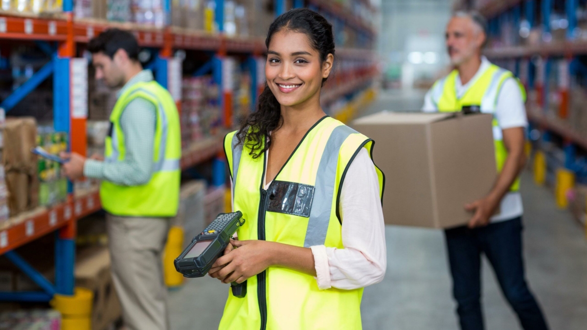 Smiling woman in a yellow safety vest holds a barcode scanner in a warehouse, while two coworkers organize and carry boxes in the background. Shelves with various items line both sides.