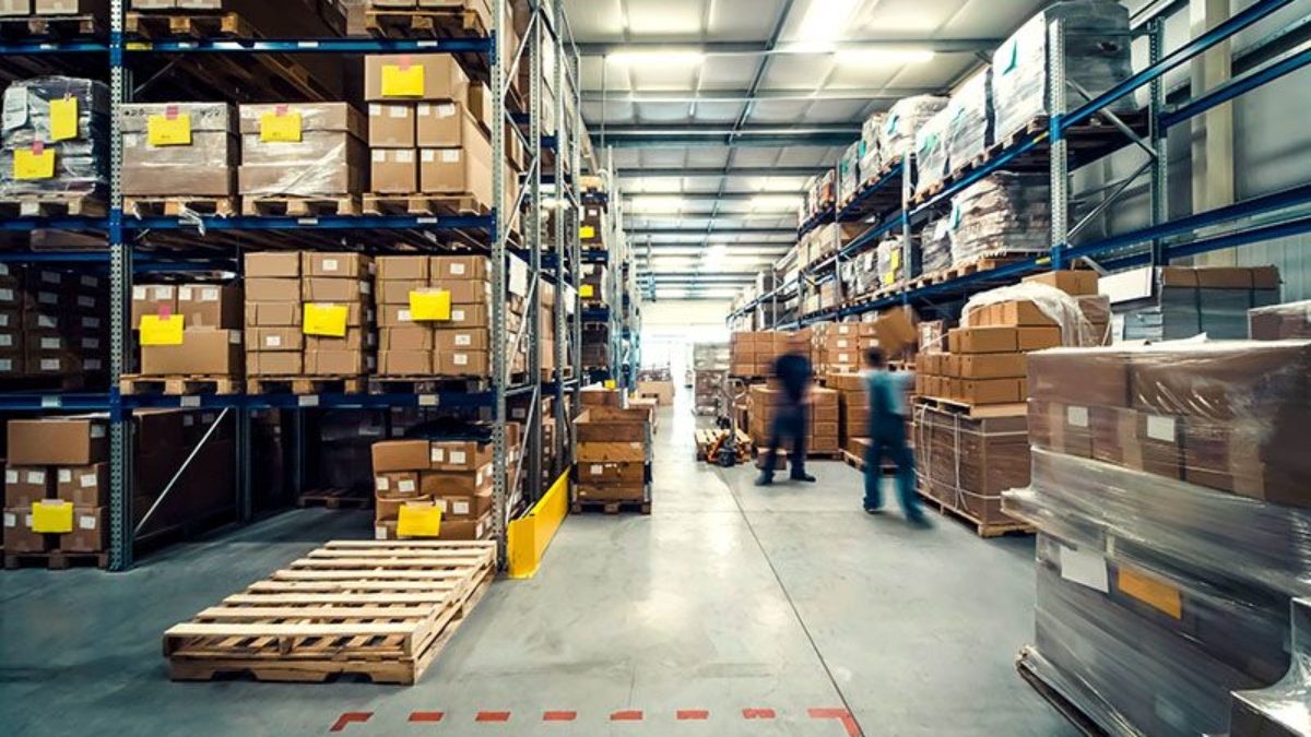 Large warehouse interior with shelves stacked high for optimal warehouse space utilization. Two blurred workers move along the aisles, and wooden pallets are visible on the floor. The brightly lit area ensures efficient operations.