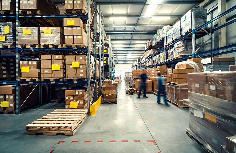 Large warehouse interior with shelves stacked high for optimal warehouse space utilization. Two blurred workers move along the aisles, and wooden pallets are visible on the floor. The brightly lit area ensures efficient operations.