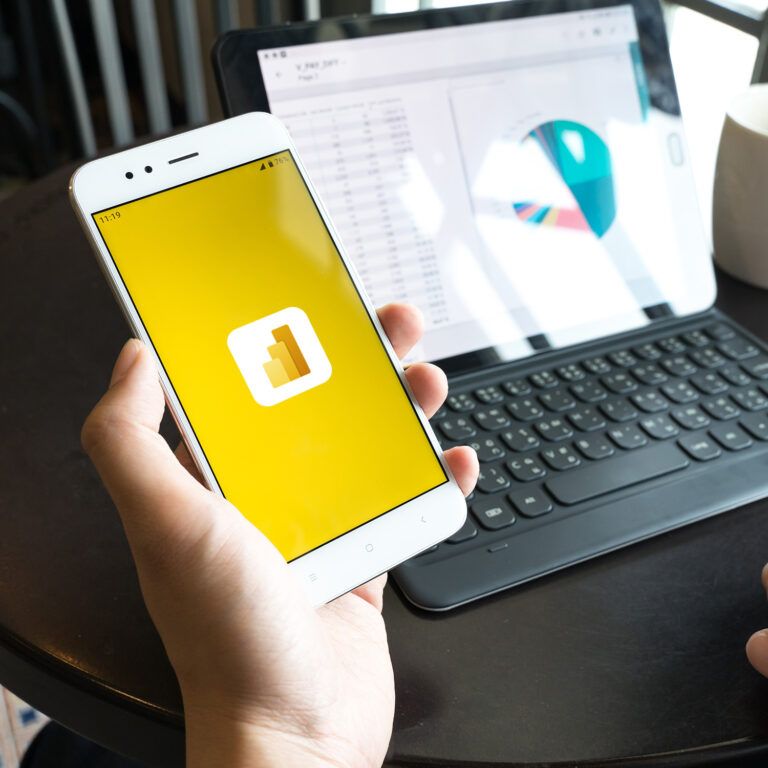 A person holds a smartphone displaying the Microsoft Power BI logo, while sitting at a round table with a tablet showing a colorful pie chart and a white coffee mug.