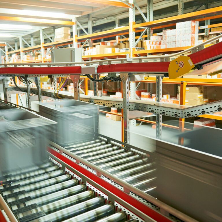 Grey storage bins move quickly along a conveyor belt system in a warehouse, with shelves stocked with various boxes and packages in the background. The motion blur highlights fast-paced automated sorting and supports efficient automation analysis.