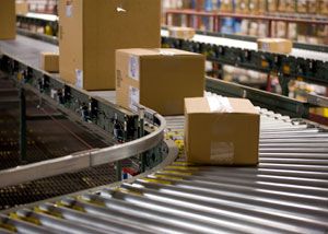 Several cardboard boxes travel along a curved conveyor belt in a warehouse or distribution center, surrounded by shelves stocked with other items.
