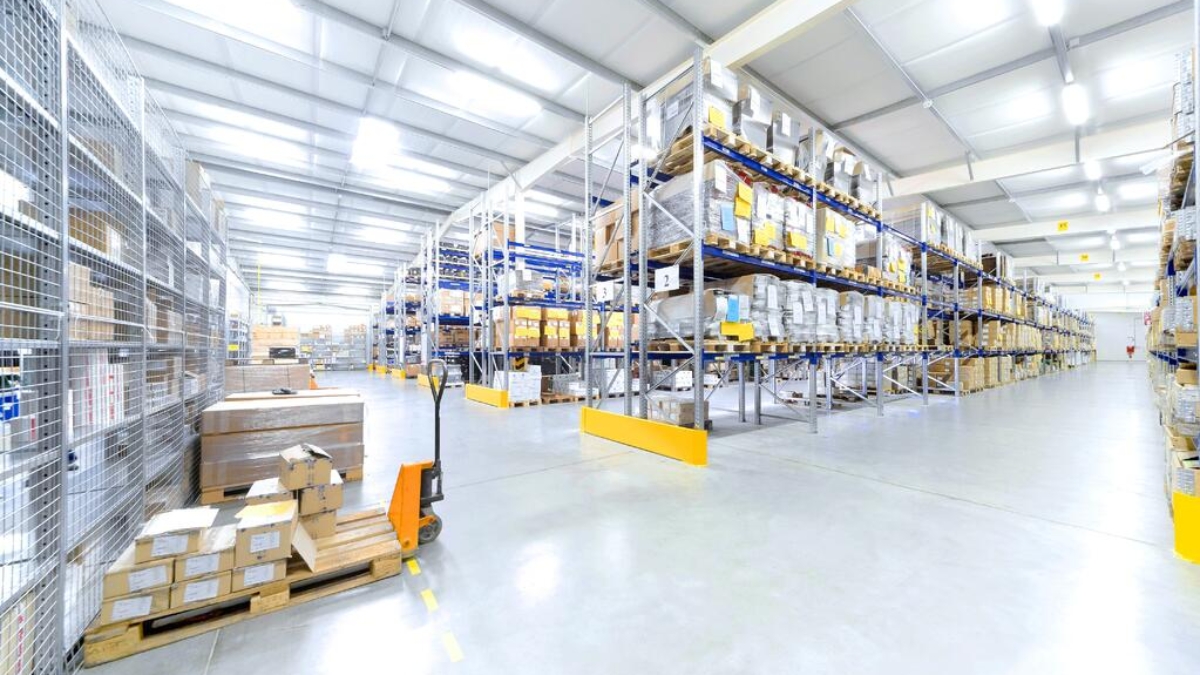 Spacious warehouse with high shelves filled with boxes and goods. Pallets and a hand pallet truck are visible on the polished concrete floor under bright overhead lighting.