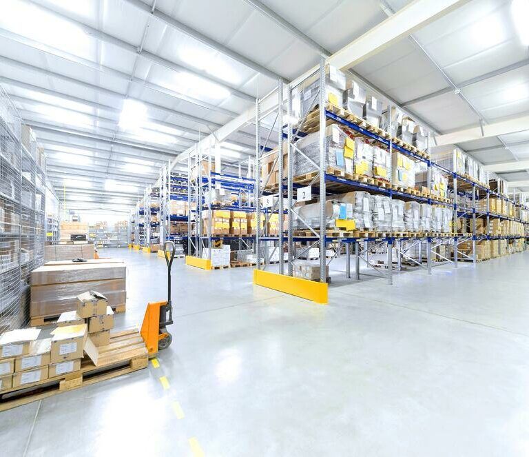 Spacious warehouse with high shelves filled with boxes and goods. Pallets and a hand pallet truck are visible on the polished concrete floor under bright overhead lighting.