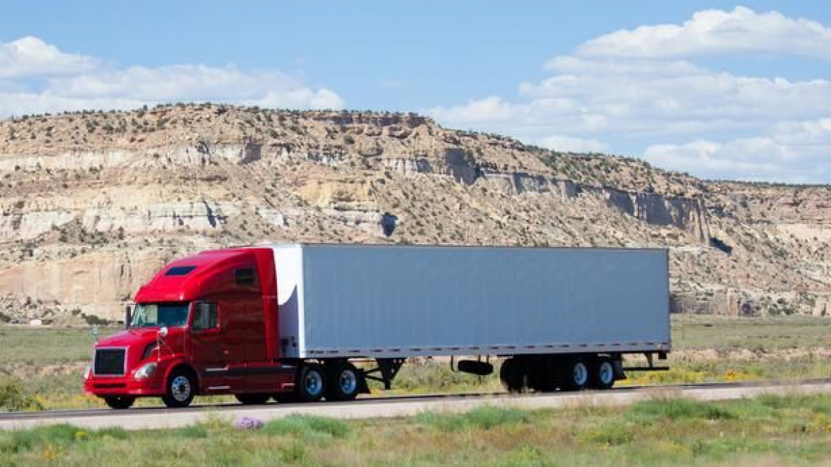 A red semi-truck with a white and gray trailer drives along a highway, with rocky, tan cliffs and a blue sky with scattered clouds in the background.