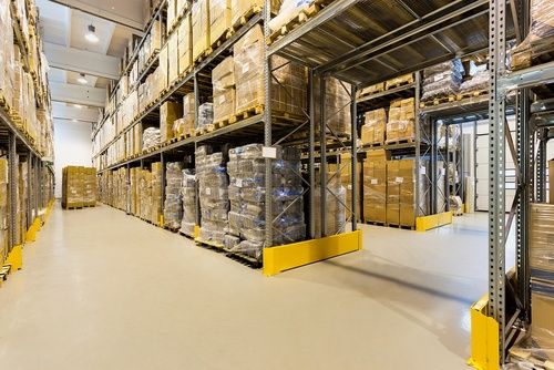 A large warehouse interior with tall metal shelves stacked with cardboard boxes and pallets. The floor is clean, and there is ample lighting. The shelves line both sides of a wide aisle.
