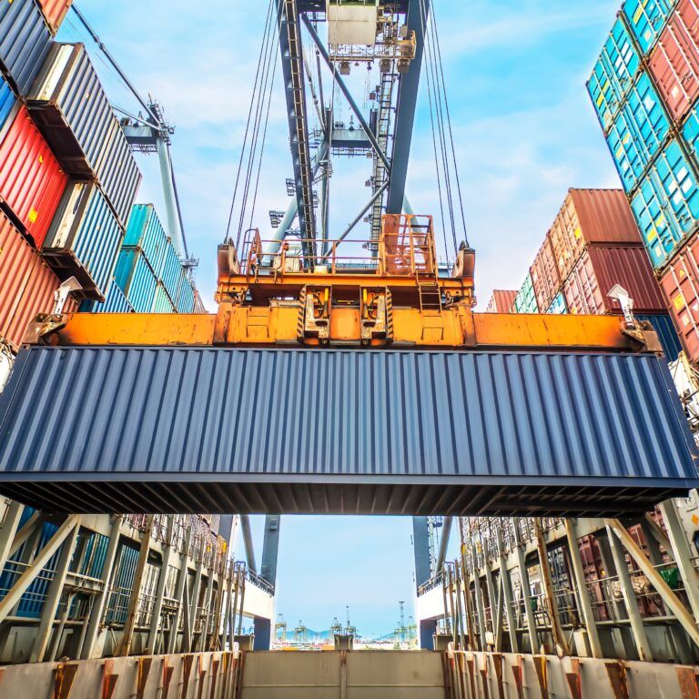 A large crane lifts a blue shipping container above a cargo ship, surrounded by stacks of colorful containers under a bright sky at a busy port.