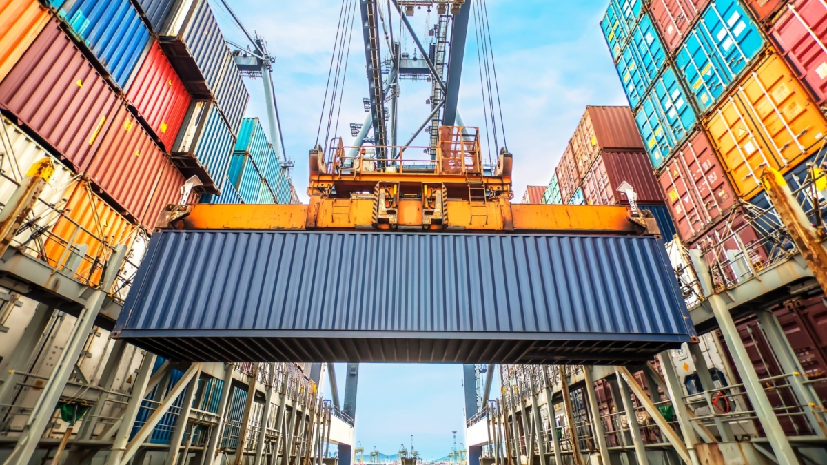 A large crane lifts a blue shipping container among stacks of colorful containers at a busy port under a blue sky.
