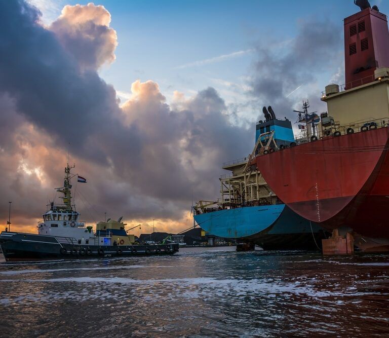 A tugboat moves near two large docked cargo ships at sunset, with dramatic clouds and golden light reflected on the water.