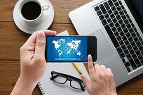 A person holds a smartphone displaying a world map with network icons. The phone is above a notepad, eyeglasses, and a keyboard, with a cup of coffee nearby on a wooden desk.