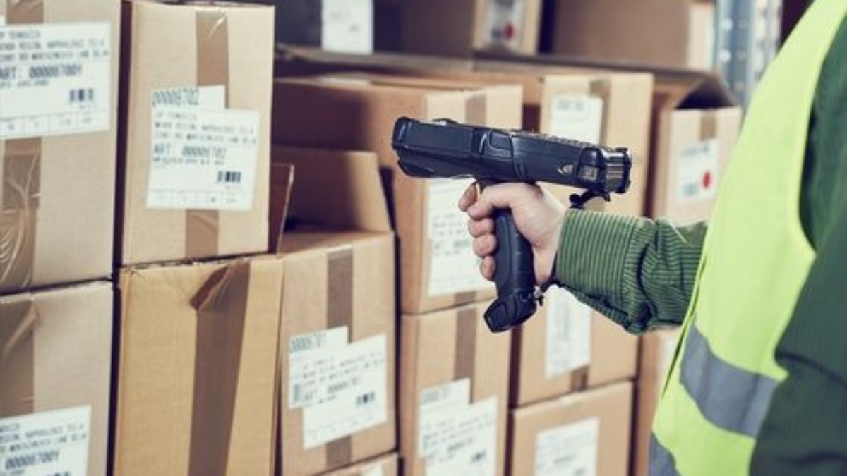 A person wearing a yellow safety vest scans barcodes on stacked cardboard boxes in a warehouse using a handheld barcode scanner.