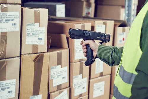 A person wearing a yellow safety vest scans barcodes on stacked cardboard boxes in a warehouse using a handheld barcode scanner.