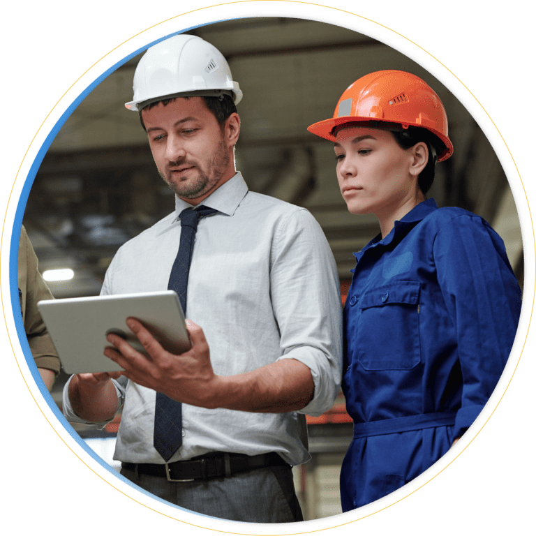 Two workers wearing safety helmets and work attire stand together. Demonstrating Business Continuity Solutions, the man in a white helmet and tie holds a tablet as the woman in an orange helmet and blue coveralls looks on attentively.