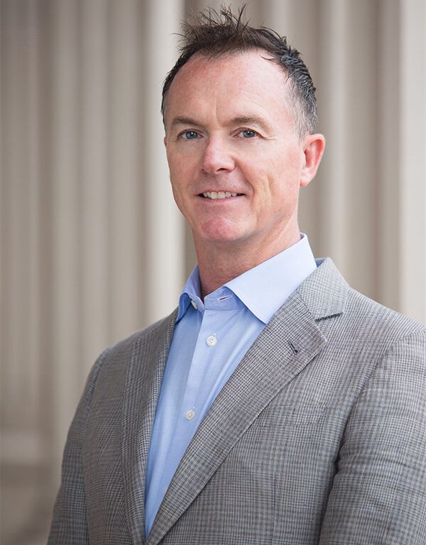 A man with short brown hair, wearing a light blue dress shirt and a gray checked blazer, stands in front of a blurred background with vertical columns. He is smiling slightly and looking at the camera, exuding quiet confidence and leadership.