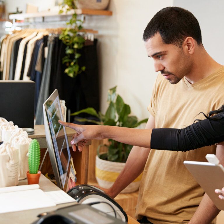 Two people stand at a retail store counter, one pointing at a computer screen while the other holds a tablet—demonstrating retail management systems. Shelves with clothing and various products are visible in the background.