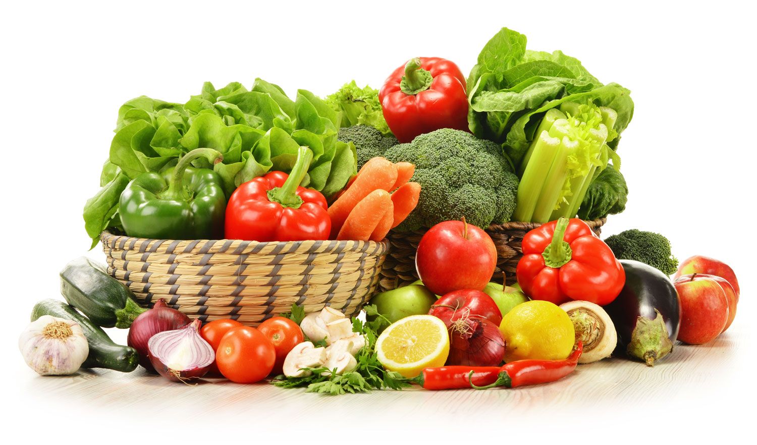 A variety of fresh vegetables and fruits, including lettuce, bell peppers, carrots, broccoli, tomatoes, onions, garlic, lemon, chili peppers, eggplant, and apples, arranged in baskets and on a white surface.