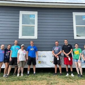 A group of nine people stand smiling in front of a gray house with two windows; some wear athletic clothes, a few wear hats. A banner with various logos hangs on the wall behind them, highlighting their commitment to community impact.