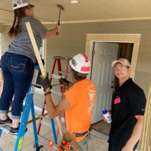 Three people work on a porch, showcasing community impact: a woman on a ladder hammers the ceiling, a man in an orange shirt and hard hat assists, while another man in a cap smiles at the camera, holding a water bottle.