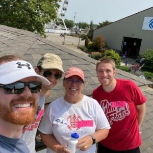 Four smiling people pose together on a rooftop, capturing a moment of community impact with a large Ferris wheel, trees, and buildings in the background. Three wear hats and sunglasses, and one holds a water bottle on this sunny day.