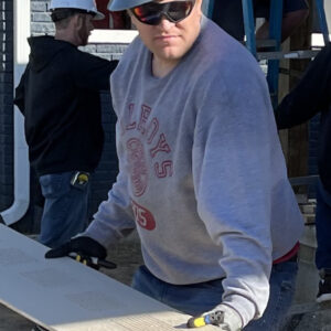 A person wearing a gray hard hat with a pink label, sunglasses, gloves, and a gray sweatshirt holds a long board at a construction site, contributing to community impact. Other workers and a ladder are visible in the background.