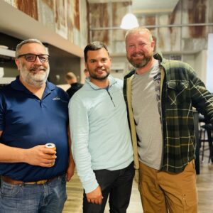 Three men stand close together, smiling at the camera in a casual indoor setting with rustic corrugated metal walls and warm lighting. One holds a drink can; they appear relaxed and friendly, reflecting the spirit of community impact.