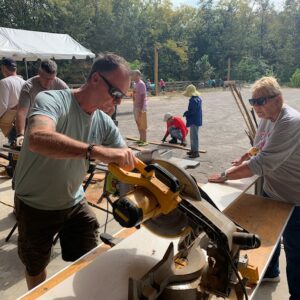 A man uses a circular saw to cut wood on a workbench outdoors, while a woman holds the wood steady. Their teamwork, along with others working on projects in the background, highlights positive community impact under the canopy and open area.